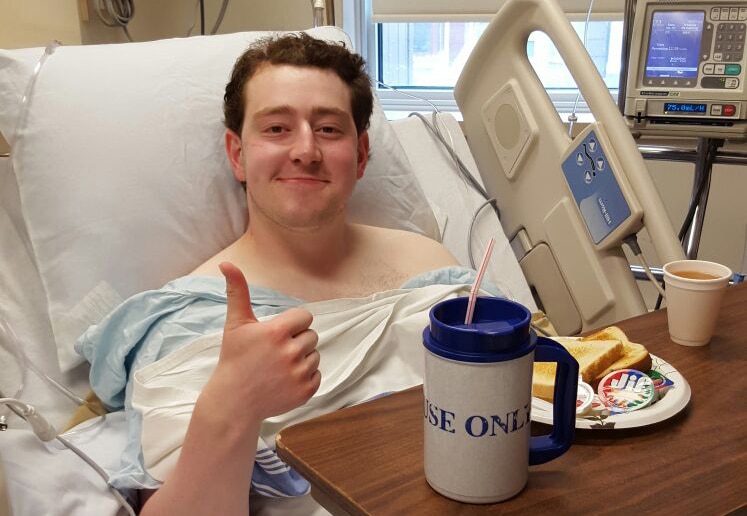 A man sitting in a hospital bed. He has a meal in front of him and is smiling, giving the thumbs up sign.