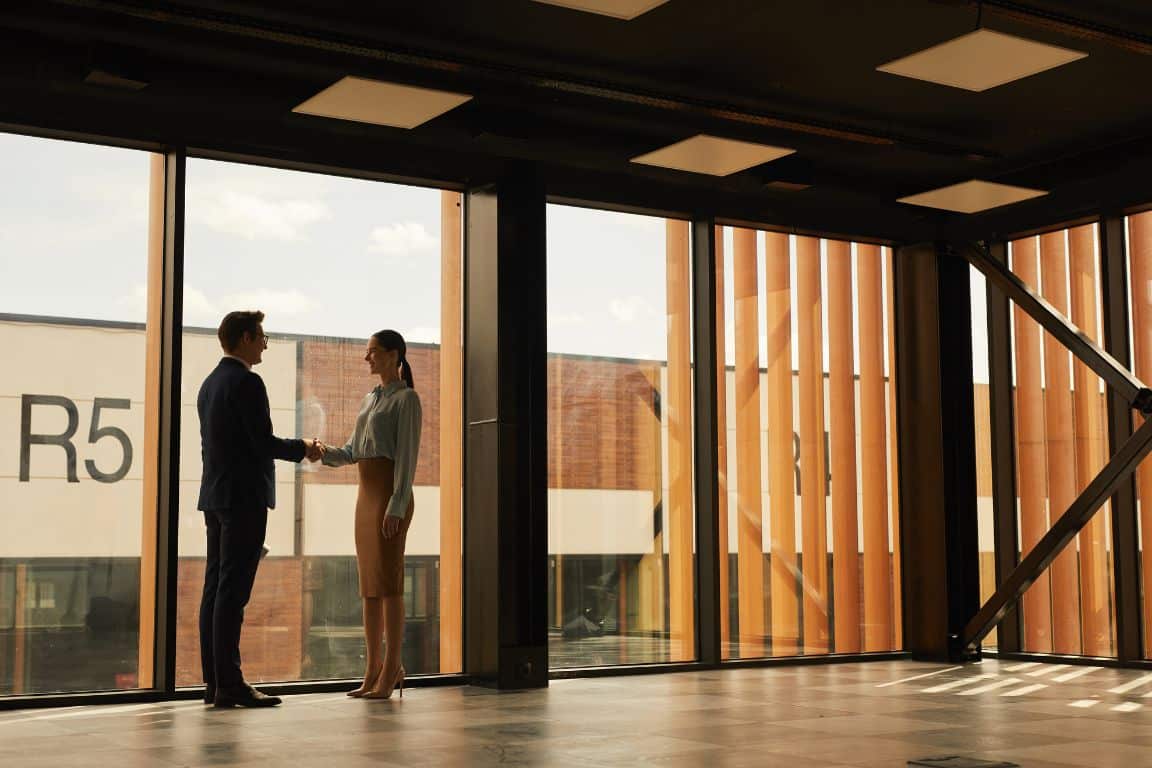Two people in business attire shaking hands in a modern, spacious office with large windows.